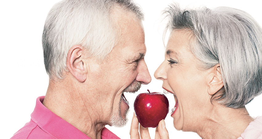Close up of an older couple facing each other and biting into an apple with beautiful and functional teeth