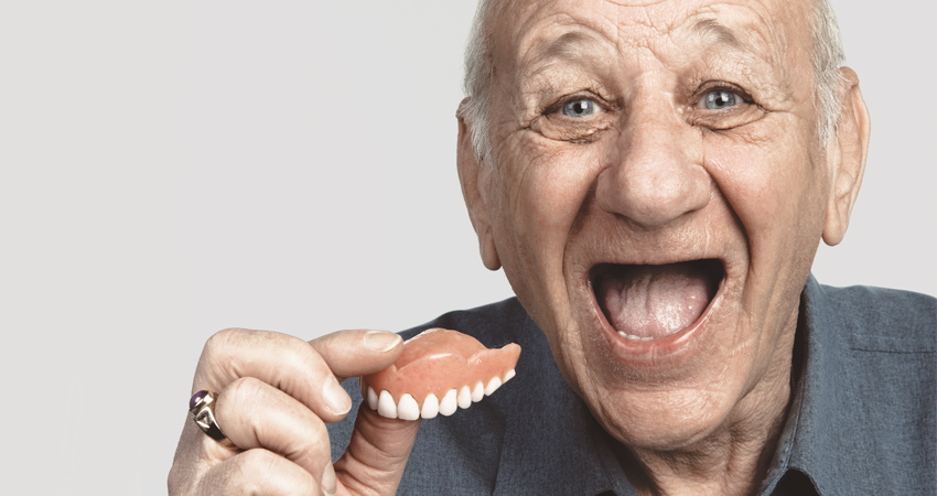 An older man who is laughing and holding his upper denture in his hand